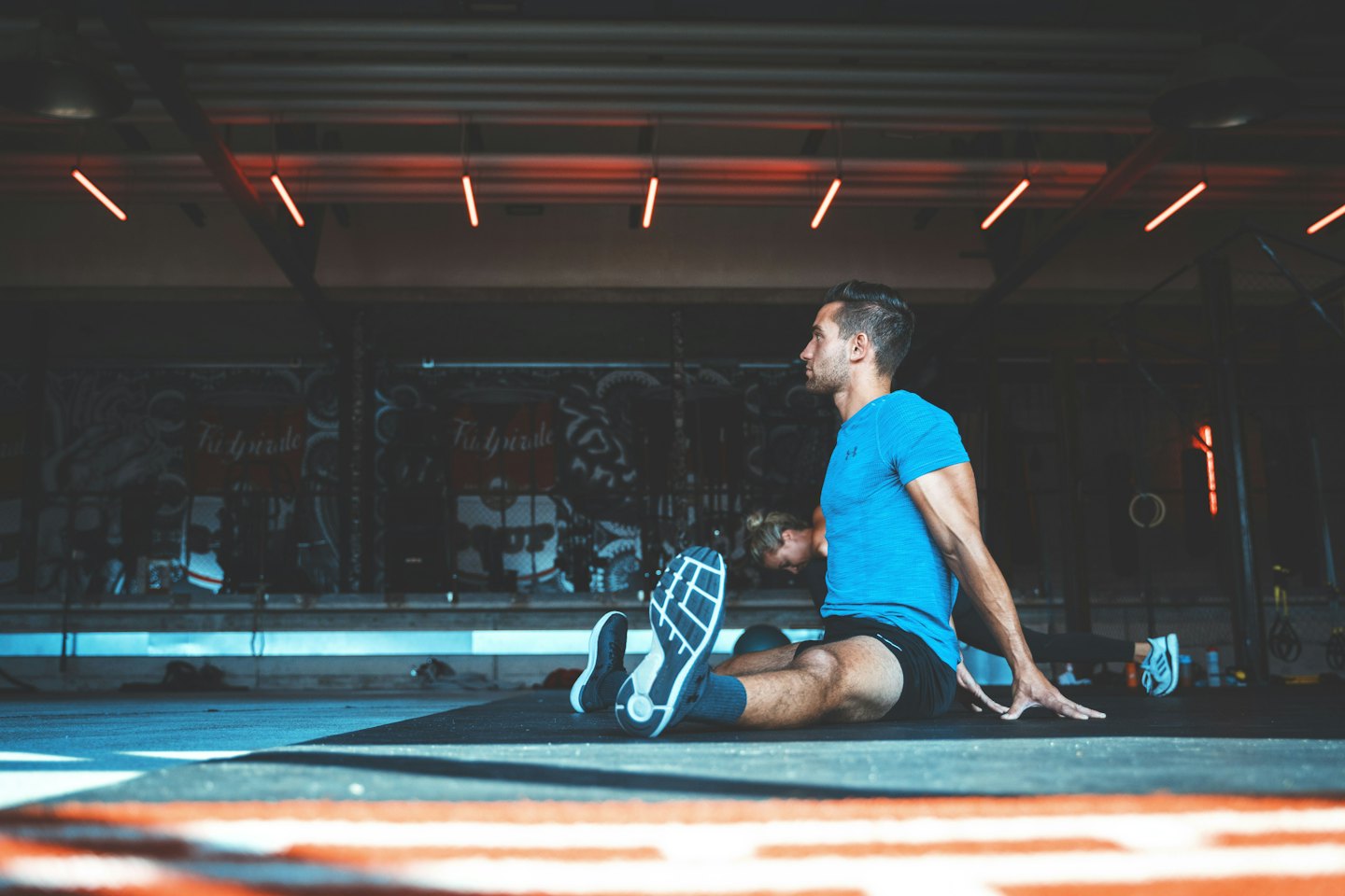 Man in blue shirt stretches on gym floor under neon lights.